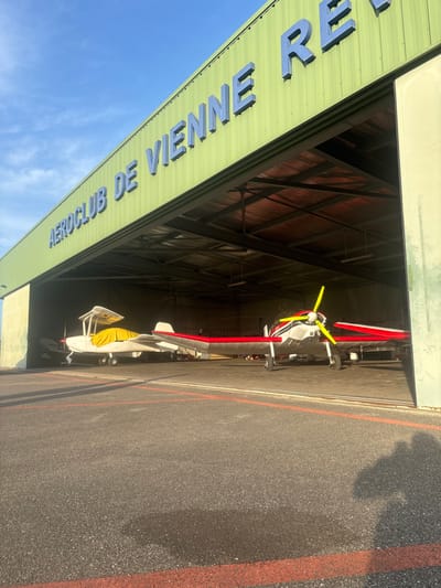 This image depicts a wide exterior view of an aircraft hangar at the Vienne-Reventin Airport (LFHR) in Chonas-l'Amballan, France, under a clear blue sky. The hangar is constructed of light-green corrugated metal, featuring large blue three-dimensional lettering on its upper facade that reads "AEROCLUB DE VIENNE REVENTIN" (partially visible). A wide, open bay reveals the interior of the hangar.
Inside, two light aircraft are visible. To the left, a white aircraft, possibly a biplane or high-wing design, is positioned deeper within the hangar, partially covered by a yellow tarp over its forward fuselage and cockpit area. To the right, a single-engine, low-wing aircraft with a white fuselage, red wing accents, and a distinctive yellow three-bladed propeller is positioned closer to the hangar's threshold, oriented with its nose facing outward. The ground in front of the hangar is an asphalt tarmac, marked with thin red lines. The scene is illuminated by direct sunlight, casting distinct shadows across the tarmac, including shadows consistent with a person or object to the right, outside the frame. No individuals are visibly present in the frame.