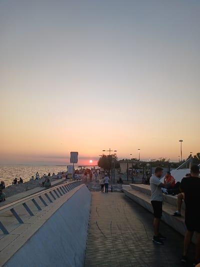 The image depicts a waterfront promenade in Karşıyaka, Turkey, at sunset. The sun, a distinct orange disc, is positioned low on the western horizon over Izmir Bay, casting an amber glow across the water and sky. The sky transitions from a pale blue at the top to shades of orange and pink near the horizon.
In the left foreground, a modern, sloped, light-colored concrete barrier with horizontal wooden slats forms a divide between a dark wooden pedestrian walkway and the rocky shoreline. To the right, a series of tiered, light-colored concrete benches provide seating. Two males stand in the immediate right foreground: one in a grey t-shirt and dark shorts, facing left; the other, partially obscured, in a black t-shirt. A third male in a red shirt is seated on the tiered benches behind them.
The midground features a wide pedestrian promenade, appearing to be paved with dark material. Approximately 50-70 individuals are present on the promenade and adjacent areas. Many are walking, while others are seated or standing on the rocky shoreline to the left, observing the sunset. Multiple streetlights and sparse trees are visible along the right side of the promenade. The water surface appears calm with subtle ripples, reflecting the sunset light. The scene represents an active urban leisure environment during twilight.