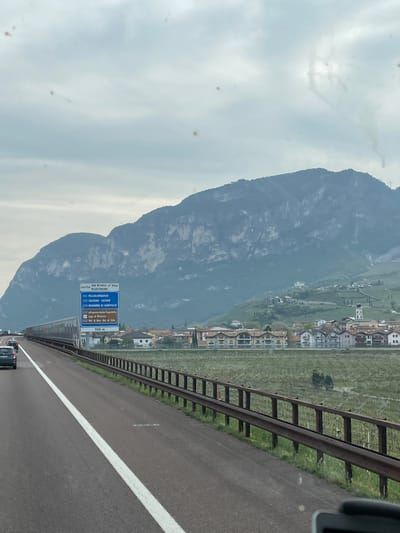 The image shows a view from a vehicle traveling on a highway near Nave San Rocco, Italy. In the background, a large mountain rises to the left of the frame. A town with closely packed buildings is situated between the mountain and the highway. A metal guardrail runs parallel to the highway. Road markings are visible on the asphalt. A blue and brown road sign indicates exits for various locations including San Michele all'Adige, Mezzocorona, Mezzolombardo, Salorno Salurn, Madonna di Campiglio, Altopiano della Paganella, Lago di Molveno, and Val di Non-Val di Sole, with a distance of 700 meters. Several cars are visible in the distance. The sky is overcast, and the overall lighting is subdued.