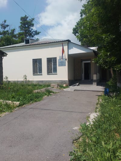 The image shows a small, cream-colored, single-story building with a metal roof, located in Nor Hachn, Armenia. The building has two windows with decorative metal bars. An Armenian flag is mounted on the exterior wall near the entrance. The entrance is a doorway with steps leading up to it. The building is surrounded by overgrown grass and trees. There is a paved pathway leading to the entrance. The sky is blue with some clouds.