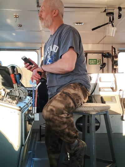 The image depicts a man sitting on a stool inside a ship's bridge. The man has gray hair and a beard. He is wearing a gray t-shirt and camouflage pants. He is holding a smartphone in his left hand. The ship's control panel is visible in the foreground, and an exit sign is visible in the background. The location of the image is not evident in the image. There is no information to conclude this is Kurdiug, Russia.