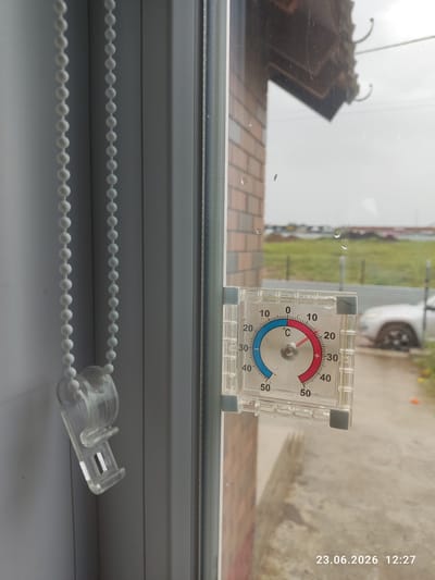 The image shows a thermometer attached to a windowpane. The thermometer indicates a temperature of approximately 15 degrees Celsius. Outside the window, a brick building, a white car, and a green field are visible. The weather appears overcast. The lower right corner of the image displays the date "23.06.2026" and time "12:27". The scene is situated in Kuyuki, Russia.