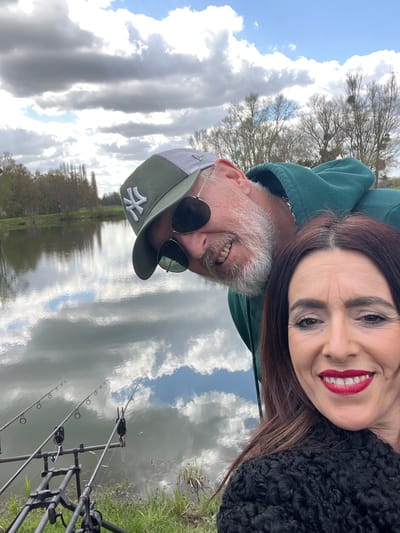 A selfie photograph taken outdoors depicts two people, likely a couple, in the context of a fishing activity. The woman is dark-haired and wearing red lipstick, while the man has a gray beard, sunglasses, and a green baseball cap with a white symbol. The photo appears to be taken at a slight angle, resulting in a tilted perspective. In the background is a body of water reflecting the sky with clouds. Fishing rods and equipment are visible near the water's edge. The location is Gavisse, France.