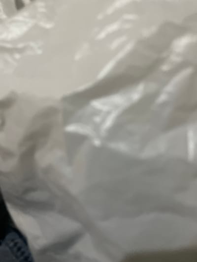 The image is a close-up, out-of-focus shot of a white, crinkled plastic bag. The texture of the plastic is the primary visual element, with highlights and shadows indicating folds and creases. A small portion of dark fabric, possibly denim, with a visible pattern of stitches, is visible in the lower left corner. The scene appears to be an indoor setting, but no specific location details within Safari, Nigeria, can be determined from the image.