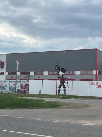The image shows a roadside scene in Staraya Kupavna, Russia. A sculpture of a robot figure made from scrap metal stands near a gray building with red trim and a white section with red text. A sign with Cyrillic text related to vehicle repair is visible. The road itself is asphalt, and the sky is cloudy.