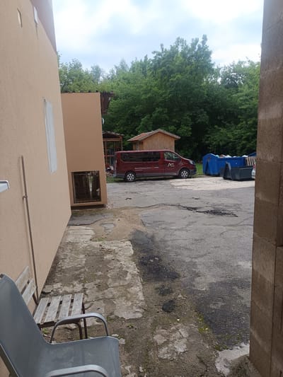 This image depicts an outdoor scene in Milovice, Czechia. In the foreground, a light-colored building wall runs along the left side of the frame, with a metal rod extending vertically. A grey metal chair with a mesh seat and back is positioned in front of a rustic wooden bench. The ground consists of cracked concrete and dirt. In the mid-ground, a maroon van with the text "AUTO licíny" and "mobility" is parked on an asphalt surface. Adjacent to the van are two large blue dumpster bins and a dark grey skip. A small wooden structure with a peaked roof is visible behind the van. The background is dominated by a dense line of lush green trees under a cloudy sky. The overall impression is of an industrial or utilitarian outdoor area, likely on the outskirts of a residential area or within a commercial property.