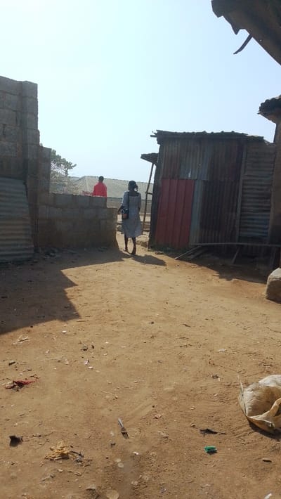 The scene depicts an unpaved outdoor area in Idogwari, Nigeria, under clear, bright daylight. The ground consists of light brown packed dirt with scattered debris, including a metal fork, a crumpled piece of light-colored fabric, and a green plastic cap. Distinct shadows are cast towards the lower left, indicating direct sunlight from the upper right.
In the mid-ground, a person, identified as female by attire and hair, is walking away from the viewer, positioned centrally on the path. She wears a light-colored, possibly patterned dress that extends below her knees, and carries a dark bag on her right shoulder. Her hair is dark and appears to be styled in braids.
To the left, a partial structure composed of grey concrete blocks forms a low wall or entrance, with corrugated metal sheeting visible directly behind it. In the background, positioned beyond this concrete wall, another individual wearing a bright red top is partially visible from the chest upwards.
To the right, a weathered structure features rusted corrugated metal siding and a reddish-brown flat panel door. Wooden supports or beams are visible at its base. An overhead section of a similar, structurally irregular building with a corrugated roof is partially visible in the upper right corner of the frame.