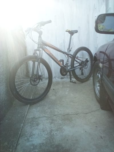 The media file shows a bicycle leaning against a wall, parked next to a car. The bicycle is a mountain bike with orange and grey accents on the frame. It has disc brakes on the front wheel. The seat is black and has "AZIMUT" written on it. The bicycle is positioned on a concrete surface, which appears to be cracked in places. A car, possibly a dark red sedan, is parked to the right of the bicycle. A side mirror of the car is visible, reflecting the sky and some greenery. The scene is lit by bright sunlight coming from the left, creating a lens flare effect and casting a strong shadow from the bicycle. The wall behind the bicycle appears to be made of concrete blocks. The overall impression is that of an outdoor setting, perhaps a driveway or a side yard. There are no people visible in the image. The time of day appears to be daytime, given the strong sunlight. The location context is Grigoriopol, Moldova.
