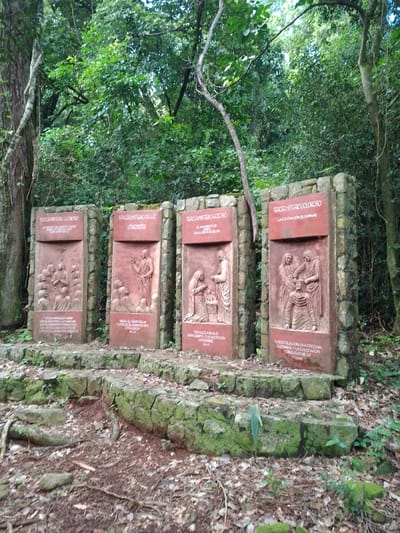Four reddish-brown stone monoliths stand on a moss-covered stone base. Each monolith has a raised carving and text. The text on the first monolith reads "TERCER MISTERIO GLORIOSO". The text on the second monolith reads "TERCER MISTERIO DE LUZ". The text on the third monolith reads "TERCER MISTERIO GOZOSO". The text on the fourth monolith reads "TERCER MISTERIO DOLOROSO". The monoliths are located in a wooded area. The location of this photograph is likely Villarrica, Paraguay due to the language of the text.