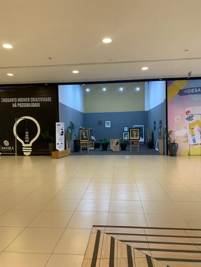 This image depicts an indoor scene within Camará Shopping, located in Camaragibe, Brazil. The setting is a clean, well-lit shopping mall corridor featuring light-colored tiled flooring, with a section of decorative wood-patterned tiles in the foreground right. Recessed ceiling lights provide illumination.
On the left, a large black banner is present, displaying the white Portuguese text "ENQUANTO HOUVER CRIATIVIDADE HÁ POSSIBILIDADE" (While there is creativity, there is possibility) and a prominent white outline of a lightbulb containing the text "TIRE SUA FOTO AQUI" (Take your photo here). The "CAMARÁ SHOPPING" logo is visible at the bottom left of this banner. A dark planter containing green foliage is positioned in front of the banner.
Directly opposite the viewer, a recessed exhibition space is visible. This area features a two-toned wall, with a darker blue-grey lower section and a lighter yellowish-grey upper section, and is illuminated by several spotlights from above. Within this space, multiple framed portraits are displayed on wooden easels and hanging on the back wall. Several dark planters with green foliage are strategically placed throughout the exhibit. Two vertical signs, one on the left and one on the right within the exhibit, partially read "EXPO. DA ARTE FOTO CAMARA Shopping," indicating a photography or art exhibition by the shopping center.
To the far right, a large, colorful banner with yellow, purple, and blue tones is visible. It features the partial text "#DESA" and illustrations of a person interacting with a mobile phone. Another dark planter with foliage is positioned in front of this banner. No individuals are present in the visible area of the image.