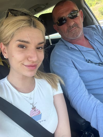 Two individuals, an older man and a younger woman, are captured in a selfie-style photograph taken inside a moving vehicle. The image is oriented vertically, with both subjects looking towards the camera.

The man, positioned in the upper part of the frame, is wearing a light blue collared shirt and dark sunglasses. He has short, greying hair and stubble, and a pair of eyeglasses on a black lanyard is visible around his neck. He displays a relaxed expression with a slight smirk.

The woman, in the foreground, has blonde hair and a gentle smile. She is wearing a white t-shirt with a seatbelt diagonally across her chest. A silver multi-strand necklace, one featuring a small red and silver cross charm, adorns her neck. Her t-shirt features a graphic of a pink and blue cupcake, with the partially visible text "...rry on top" next to it, likely part of "Cherry on top."

The setting is an interior of a car or similar vehicle, with elements like the seatbelt, a car door handle, and reflections in the sunglasses confirming this. Through the windows, blurred green foliage and what appears to be a road suggest the vehicle is in motion during daylight hours. Both subjects appear relaxed and happy. The location context provided is Jeleznitsa, Bulgaria.