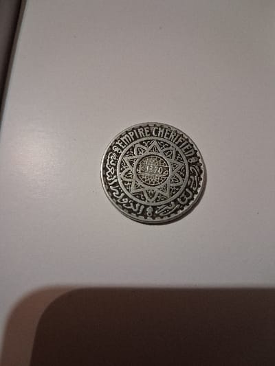 A silver-colored coin lies on a white surface. The coin features intricate designs, including a central star shape containing what appears to be a date or number, possibly "1370." Encircling this are inscriptions in both Arabic and Latin script. The Latin text reads "EMPIRE CHERIFIEN." The coin's design is complex and ornate.