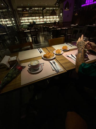 The image captures a dimly lit, intimate setting inside a restaurant or cafe, likely in Lomas de Zamora, Argentina. The scene is viewed from a slightly overhead, tilted perspective, focusing on a wooden table prepared for two.
In the upper-left of the main image, a person's arm, dressed in a green top, is visible. Their left hand, adorned with extensive tattoos including an eye design and the number "1998" on the wrist, holds a clear glass.
On the table, several items are arranged on light-colored and pink placemats. There are three white plates, each containing a golden-brown pastry, possibly a croissant. A cup of coffee with latte art rests on a matching saucer with a pink patterned rim. Cutlery sets, including forks and knives, are neatly placed on the placemats. A small wooden dispenser filled with white napkins sits near the edge. Notably, a single red rose, wrapped in cellophane, lies on the table, suggesting a special occasion or romantic gesture.
The background shows the restaurant's interior with shelving units displaying various items, and other tables and chairs, largely unoccupied. The lighting is low and atmospheric, primarily from artificial sources. A purple neon sign on a wall is visible, displaying the text "ROSE COCINA CASERA NON-STO," indicating the restaurant's name and a specialty in homemade cuisine. Reflections of the interior lights can be seen on a dark, glossy surface, likely the floor. The overall ambiance suggests an evening outing for coffee and pastries in a cozy environment.