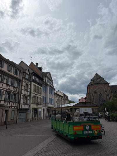 A green tourist train is parked on a cobblestone street in Colmar, France. The train is filled with passengers. In the background, there are traditional Alsatian buildings with half-timbered facades and a stone church with a pointed roof. The sky is overcast.