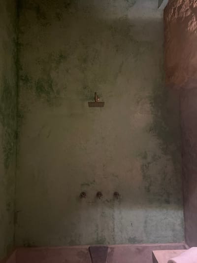 An empty, green-tinted shower room features textured walls and floor with subtle variations in color and pattern. A central, horizontal shower handle is visible on the left side, while three smaller shower heads are aligned vertically on the right. The ceiling appears slightly rocky or uneven. Part of a structure, possibly a wall or shelf, is visible on the right side. Overall, the room has a rustic or natural aesthetic.