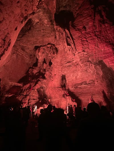 The image depicts a group of people gathered inside a cave, illuminated by red lighting. The cave walls are rough and textured, with visible rock formations and crevices. Several individuals are scattered throughout the cave, some standing and some seated on the rocky ledges. The foreground is dominated by the dark silhouettes of more people, suggesting a tour or a performance. The scene appears to be in Gollagutta, India, within a cave environment.