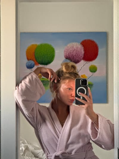 A young woman with blonde hair styled in a bun, wearing a pink terrycloth robe, is taking a mirror selfie in what appears to be a bedroom. She is holding a smartphone to capture her reflection. Behind her, a painting depicts a surreal landscape with colorful, spherical trees and a body of water under a blue sky. The room also shows a glimpse of a white, rumpled bed. The overall scene suggests a relaxed, private moment. There is no indication of the location being in Ariel, Palestinian Territories.