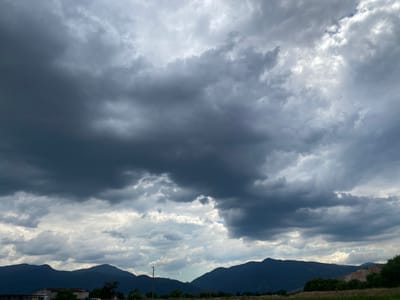 The image captures a dramatic sky over Kurtovo Konare, Bulgaria, dominated by dark, heavy storm clouds. The clouds are dense and varied in texture, with lighter patches suggesting breaks in the overcast sky. In the foreground, a range of dark, silhouetted mountains stretches across the horizon. Below the mountains, there are hints of human settlement, including a few structures that appear to be buildings, possibly agricultural or industrial, and some vegetation. Power lines are also visible, indicating infrastructure. The overall impression is one of an impending storm in a rural or semi-rural setting.