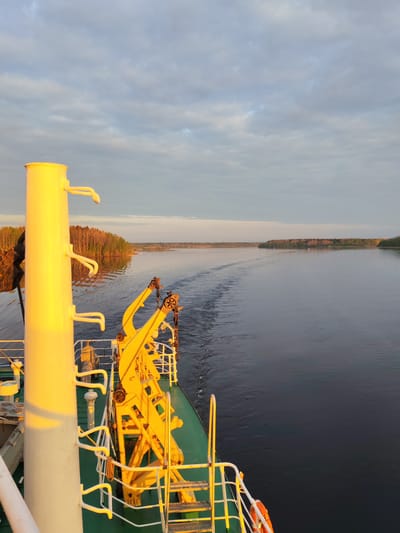 View from a ship, possibly on the Dnieper River, near Pochinok, Russia. The ship has yellow machinery and white railings. The water is smooth with a wake visible behind the ship. Trees line the distant shore. The sky is partly cloudy with the sun low, giving the ship a warm glow.