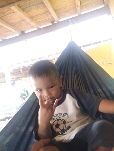 In Pompanua, Indonesia, a young boy sits in a hammock, his left arm outstretched as he makes a hand gesture. The hammock is made of dark fabric and is suspended from a wooden structure with a sloped, thatched roof. The boy wears a t-shirt with a soccer ball graphic and the word "VICTORY" printed on it. The background features a yellow wall with some indistinct markings and bright sunlight filtering through, creating a lens flare effect. The overall scene appears to be outdoors, possibly in a covered patio or an open-air dwelling.