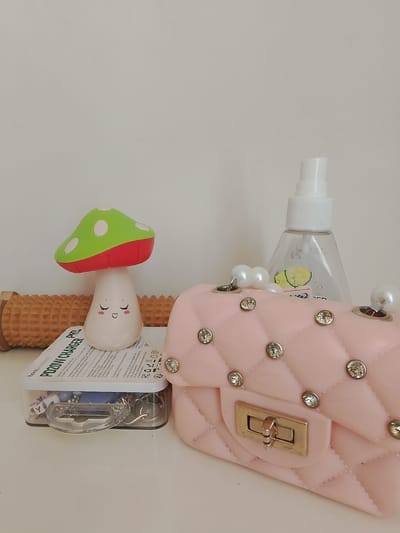This is a still life image featuring a collection of small decorative items and personal care products arranged on a white surface against a plain white background. The primary subjects are a pink quilted purse adorned with rhinestones and pearl-like beads, a small white figurine of a mushroom with a green and red cap and a smiling face, a clear spray bottle, and a wooden foot roller.
The pink purse is in the foreground, positioned on the right side of the frame, with its quilted texture and decorative hardware prominently visible. Behind it and slightly to the left, the mushroom figurine stands, appearing whimsical with its closed eyes and blushing cheeks. A clear spray bottle, possibly containing hand sanitizer or a facial mist, is situated behind the mushroom. The foot roller, a cylindrical wooden massager, is visible on the far left, partially obscured. A clear plastic container holding various small charms or beads is placed in front of the foot roller and under the mushroom figurine.
There is visible text on the packaging of the foot roller, including "hoco.accessory" and "POZINN CHARGER AC-240". The overall arrangement suggests a curated display of personal items, possibly on a vanity or shelf. There are no people visible, and the setting appears to be indoors, with neutral lighting and no indication of time of day or weather. The emotion conveyed is one of playful decoration and personal care. The location context provided is Krasnostepnovskoye, Russia.