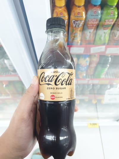 A hand is holding a bottle of Coca-Cola Zero Sugar Vanilla flavored drink in a convenience store refrigerator. The bottle is clear plastic with a black cap and a gold label. The label has "Coca-Cola" in black script, "vanila" in a smaller font above, and "ZERO SUGAR" in bold black letters below. There is also text that reads "BEBAS GULA" which translates to "sugar-free" in Indonesian. The "BARU" label indicates the product is new. The bottle has a small Halal certification logo. In the background, out of focus, are shelves stocked with various bottled drinks. The environment suggests an indoor retail setting, possibly a minimart or supermarket, with bright fluorescent lighting. The scene is likely in Proyonangg, Indonesia, given the Indonesian text on the label.