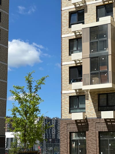 This image, taken in Znamya Oktyabrya, Russia, captures a residential building under a clear blue sky with scattered white clouds. The foreground features a young green tree with abundant leaves, partially obscuring the view of a multi-story apartment complex. The building itself is constructed with light-colored brick and features numerous windows, some with visible curtains. Air conditioning units are mounted on the exterior walls. Another apartment building, appearing taller and with a darker facade, is visible in the background, along with a street light and fencing. The scene is a snapshot of an urban residential environment on a bright, sunny day.