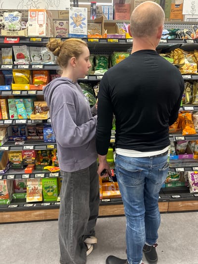 The image captures a shopping scene inside a supermarket aisle, specifically at Woolworths Toormina, Australia. A young woman with auburn hair styled in a bun, wearing a grey hooded sweatshirt and grey wide-leg jeans, stands facing away from the camera, examining a shelf of snacks. To her right, a bald man, seen from the back, is also looking at the shelves, dressed in a black long-sleeved shirt and blue jeans. Both individuals appear to be in the process of grocery shopping. The shelves are well-stocked with various packaged food items, including rice cakes, granola, chips, and other snack products, each with visible price tags. The lighting is typical of an indoor retail environment, with overhead fluorescent lights illuminating the products and aisles. The floor is a light grey concrete.