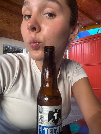 A selfie-style image depicts a young female posing with a bottle of Tecate beer in Ciudad Victoria, Mexico. The subject, positioned in the foreground, has light brown hair, brown eyes, and is wearing a white t-shirt. Her lips are puckered. A single white pearl earring is visible in her right ear. A brown glass bottle of Tecate beer, exhibiting condensation and the brand's label featuring an eagle logo and "TECATE" text, is held or positioned in front of her, obscuring her lower torso. The bottle rests on a dark, reflective surface. The background on the left reveals a thatched roof structure with wooden supports, while the right displays a bright red wall with vertical slats and an overhead blue and possibly yellow structure.