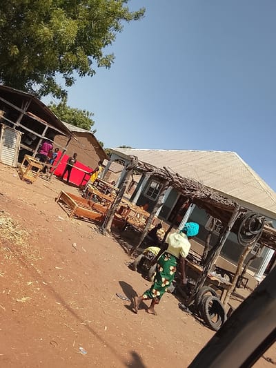 The image portrays an outdoor commercial or workshop area in Ibi, Nigeria, under a clear blue sky. A large green tree canopy is visible in the upper left quadrant. The ground throughout the scene consists of reddish-brown dirt, with scattered wood shavings and debris.
On the left, an open-sided structure with a wooden frame and corrugated metal elements functions as a carpentry workshop. Inside, a person in a purple garment is visible. Outside this structure, several wooden furniture items, including tables and bed frames in various stages of completion, are displayed. In front of this area, a person in a black shirt stands next to a large, bright red rectangular piece of fabric, possibly preparing or moving it. Another individual in a yellow shirt is also present nearby.
In the mid-ground, a woman walks from right to left, facing away from the viewer. She wears a light yellow short-sleeved top, a green skirt patterned with lighter motifs, and a light blue head covering. She is wearing sandals. Her shadow is cast on the dirt path ahead of her.
To the right, a building with a light grey corrugated metal roof features a rustic, open-air structure in front of it, constructed from wooden posts and interwoven branches. Under this canopy, multiple vehicle tires are stacked and displayed, indicating a tire sales or repair point. A partially visible individual is seated under this awning.
The extreme lower right foreground shows a dark, curved object, consistent with a vehicle tire or fender, suggesting the photograph was taken from a vehicle or immediately adjacent to one. A small, white rectangular object lies on the dirt ground in the lower-middle left.