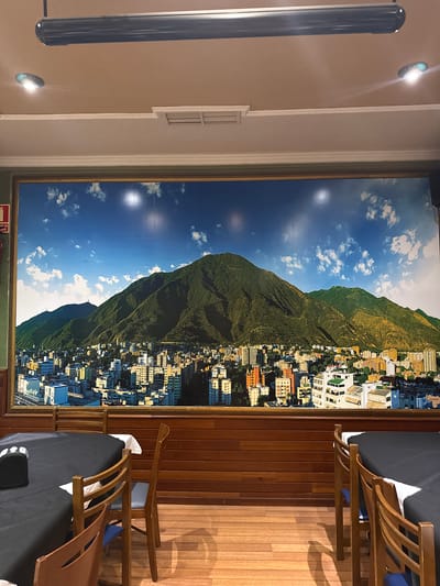 The image depicts an indoor dining area with a large mural of a city and mountains covering the back wall. The mural shows a dense urban landscape with numerous buildings of varying heights and colors, set against the backdrop of lush green mountains under a bright blue sky with scattered white clouds. The mural is framed by a golden border. Below the mural, the wall is paneled with horizontal wooden planks. In the foreground, two round tables are set with black tablecloths and white napkins. Several wooden chairs with blue seats are placed around the tables. The floor is made of light-colored wooden planks. In the upper part of the image, a dark, cylindrical fixture, possibly a projector or light, is mounted on the ceiling, along with two recessed circular lights. A red fire extinguisher is partially visible on the left edge of the mural. The overall setting suggests a restaurant or dining hall. The location of Torrejón de Ardoz, Spain, is not discernible from the visual information present in the image.