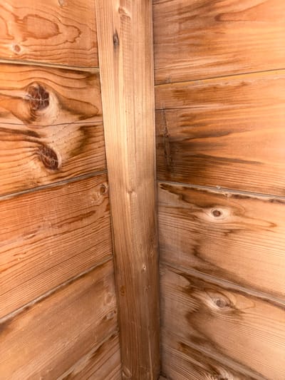 The image displays an interior corner of a wooden structure, likely a shed or an outdoor building, composed of horizontal wooden planks. A vertical wooden beam runs diagonally through the center of the image, joining two walls. The wood grain is prominent, with several knots visible in the planks. Some planks exhibit discoloration, suggesting water damage or staining. The overall lighting is dim, casting a warm, amber tone on the wood. The scene is devoid of any people, actions, or interactions. The location in Novoanninskiy, Russia, is not discernible from the visual information presented in the image.