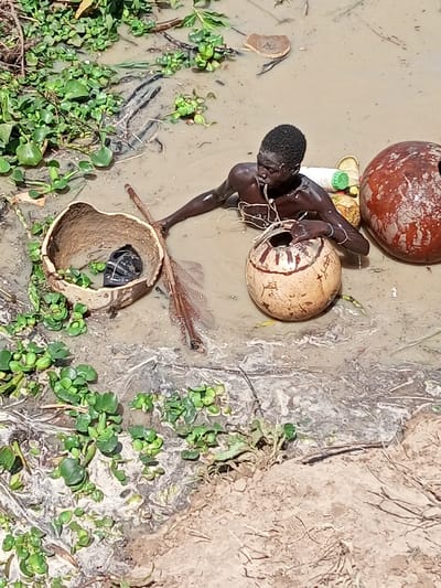 In Kokani North, Nigeria, a male individual is submerged from the chest down in light brown, muddy water. He is bare-chested, with white string visible around his neck and light-colored wristbands on both wrists. With his left hand, he holds a light brown gourd adorned with Arabic script and featuring an open top. To his right, a large, reddish-brown spherical gourd floats on the water. To his left, a split gourd, shaped like a bowl, contains a dark, wrapped object. A long wooden stick is partially submerged adjacent to the split gourd. A white plastic bottle and a green container float behind the individual, near the spherical gourd. The muddy water surface is extensively covered with green aquatic vegetation and some dark debris. A dry, earthy bank is visible in the bottom right foreground.