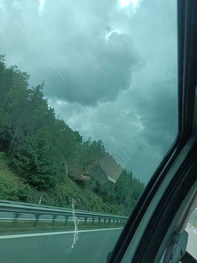 The image presents a view from inside a moving vehicle, looking outward through a tinted side window. The scene depicts a highway bordered by a steep, densely forested hillside on the left. The forest consists primarily of green deciduous trees. The highway in the lower portion of the image features a multi-tiered metal guardrail and white painted lane markings on the asphalt surface.
The sky above is heavily overcast with dark, voluminous grey and cyan-tinted clouds, suggesting stormy or impending rain conditions, with a brighter light source visible in the upper left quadrant. A dark, vertical structural element, likely a vehicle pillar or door frame, occupies the far right side of the image.
Prominent reflections are visible on the window glass. These include a clear reflection of a human hand and forearm in the mid-right section, and a distinct, stylized white, sinuous mark or reflection in the lower central part of the window. The overall greenish-blue hue of the image is consistent with viewing through tinted glass. The depicted location is a highway landscape near Hvězdonice, Czechia.