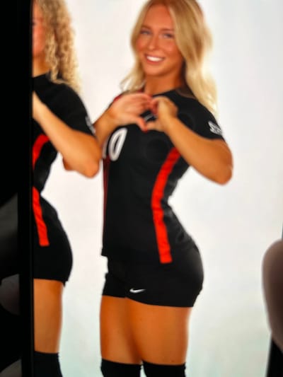 The image depicts two individuals in a studio setting within Teaneck Township, United States. A blonde female subject, positioned centrally, is smiling at the viewer while forming a heart shape with her hands. She is wearing a black short-sleeved athletic jersey with a red vertical stripe and a white number "10" on the front. Below, she wears black athletic shorts with a white Nike swoosh logo on the left thigh, and black knee-high socks or pads. To her left, a second blonde female is partially visible, wearing similar black and red athletic attire, with her left side obscured by a dark vertical element. The background is a plain white wall, indicative of a controlled photoshoot environment. The image itself appears to be a photograph of a digital display.