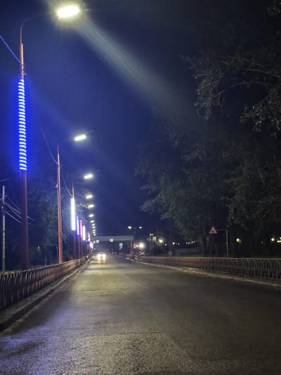 A nighttime view of an empty, wet road in Cheremkhovo, Russia, illuminated by streetlights. On the left, a series of tall poles line the road, some with bright blue LED lights and others with white street lamps. The road curves gently into the distance, where a car is visible approaching. Trees with dark foliage overhang the right side of the road, and a metal fence runs along the left side. Several traffic signs are visible, including one with a triangular warning symbol. The overall atmosphere is dark and quiet.