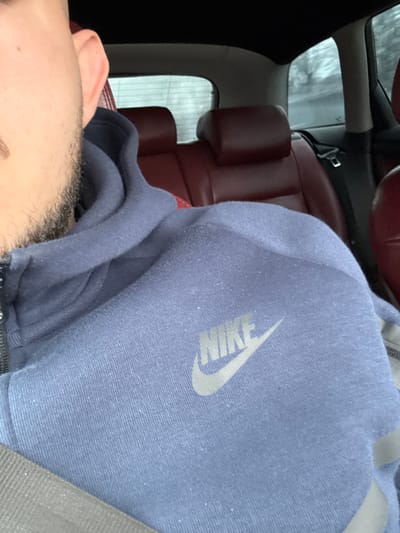The scene is set inside a vehicle, located in Malinovo, Bulgaria. A male individual occupies the foreground, with only the right side of his head (ear and lower cheek with dark stubble) and upper torso visible. He is wearing a dark blue, possibly faded, hooded sweatshirt with a zippered front. A light grey "NIKE" logo is prominently displayed on the upper left chest area of the sweatshirt. A grey seatbelt strap diagonally crosses from the lower right to the upper left across his chest. The vehicle's interior features dark red leather upholstery for the rear seats, with two headrests clearly visible. The car's headliner is black. Through the rear window, a blurred outdoor environment is discernible, characterized by indistinct trees or vegetation, suggesting motion or an overcast day. No direct interactions are depicted.