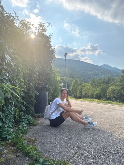 A woman is sitting on a paved road, leaning back with her arms behind her. She is wearing a white t-shirt, black shorts, and light blue sneakers. She has a white backpack on and is holding a phone in her hand. She is smiling and appears relaxed. The scene is a roadside with lush green trees and mountains in the background. The sun is shining, and the sky is blue with some clouds. The image was taken in Calceranica al Lago, Italy, likely in the late afternoon given the golden light. There is a trash can next to her. The road appears to be a rural road, possibly on a mountain pass given the elevation and foliage.