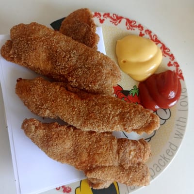 A plate of golden-brown fried food, possibly chicken tenders or fish sticks, rests on a white surface. The food is arranged in a stack, with three pieces clearly visible on top and what appears to be more beneath. Beside the food, on a decorative plate featuring red strawberries and white Mickey Mouse ears, are two dollops of condiments: one yellow, likely mustard or cheese sauce, and the other red, resembling ketchup. A white piece of paper is tucked beneath some of the food items. The plate has text around its rim that partially reads "packed with love" and contains the silhouette of a Mickey Mouse ear. The lighting suggests daylight. The scene is a close-up shot of a meal, likely a snack or part of a larger meal, in a casual setting.
