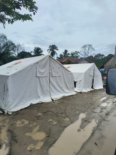 Two white tents sit in a muddy area. The ground is wet and filled with puddles. Buildings, including a house with a rusty roof, are in the background. Trees surround the area, and the sky is cloudy. A partial image of a vehicle appears in the right foreground. The tents seem to be temporary shelters in a potentially disaster-stricken area.