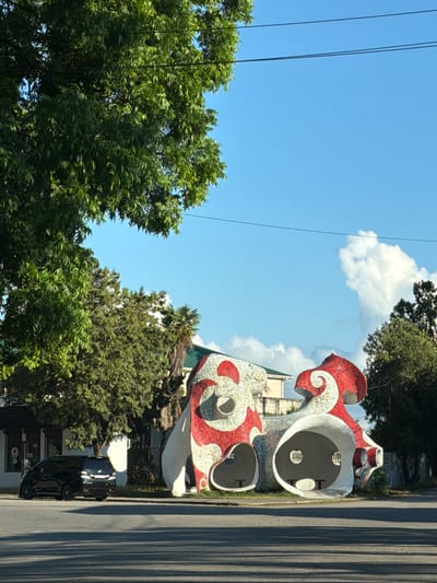 In Inkiti, Abkhazia, a mosaic-covered sculptural structure with white and red swirling patterns is situated on the side of a paved road. The sculpture features large, hollow openings, suggesting it might be a bus stop or an artistic installation. A black minivan is parked on the street to the left of the sculpture. Lush green trees with dense foliage partially obscure the view of buildings in the background. The sky above is clear blue with scattered white clouds. The scene is captured in daylight, with shadows cast on the road from the trees and the structure.