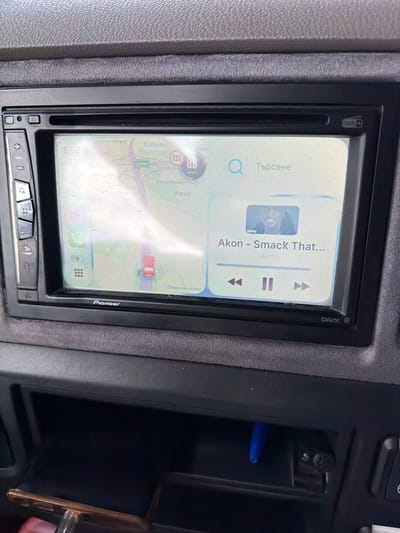 The image shows a Pioneer car stereo head unit integrated into a car's dashboard, likely in Kortrijk, Belgium. The touchscreen display presents a split interface, featuring both music playback and a navigation map.
On the upper part of the screen, a music player is active, displaying "Akon - Smack That..." by Akon, accompanied by a small album art thumbnail and standard playback controls (play/pause, skip forward/backward). To the left, a search bar shows the word "Търсене," which is Bulgarian for "Search."
The lower section of the screen displays a detailed navigation map with various street names such as "Doomanstraat" and "Delacroixstraat," and towns including "Aalbeke," "Klarenhoek," and "Marke," consistent with the Kortrijk region. The E403 highway is visible, along with a speed limit indicator showing "120 км/ч" (120 km/h). A red car icon marks the current position on the map. In the top left corner of the screen, the time "8:58" and a "Full 5G" signal indicator are visible.
The head unit itself has "Pioneer" branding on its bottom right bezel, and "dab" (Digital Audio Broadcasting) and "DIVX" logos on its top left and right bezels, respectively. Physical buttons below the screen include controls for "VOL" (volume) and "DISP" (display).
To the right of the head unit, an open storage compartment contains a blue pen. A darker compartment below it holds another object. The dashboard surrounding the unit is textured and appears to be dark grey.