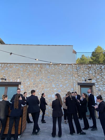 In the city of Bullas, Spain, a group of individuals, appearing to be event staff or hospitality workers, are gathered outdoors near a stone building. They are all dressed in formal black attire, predominantly black suits and trousers. The setting is likely a venue, possibly a winery or a reception area, given the rustic stone wall of the building and the outdoor lighting stringed overhead.

The individuals are arranged in small clusters, with some engaged in conversation. One person is looking up at the sky, while others are interacting with each other or facing away from the camera. A wooden bar or counter is visible to the left, suggesting a service or hospitality function. The ground is paved, and the overall atmosphere appears to be a relaxed gathering before or during an event. The sky above is clear and blue, indicating daytime. The architecture of the building features a light-colored facade above the stone, with a dark-railed balcony or walkway visible on the upper level. Trees are visible in the background, suggesting a slightly rural or landscaped environment.