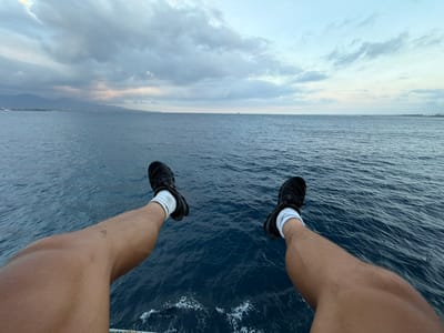 A first-person perspective shot shows a pair of legs and feet dangling over the edge of a boat or structure, with the deep blue ocean stretching out to the horizon. The legs are tanned and bare, with black athletic shoes and white socks visible. The sea is calm, with small ripples and white foam visible where waves are breaking. In the distance, the coastline of Melaya, Indonesia, can be seen, with mountains and a hazy sky. The sky is overcast, with dramatic clouds in shades of gray and hints of pink and yellow as the sun sets or rises. The overall scene conveys a sense of freedom and tranquility.