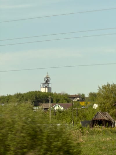 The image shows a blurred landscape near Совхозный, Russia. Power lines run across the left side of the frame. In the distance, a communication tower is visible. Vegetation and buildings are on the right, partially obscured by motion blur.