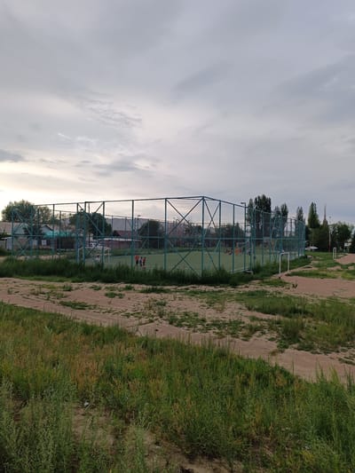 An outdoor soccer field is enclosed by a tall, green metal fence. Several children are visible on the field, some playing with a ball. The field is a light green artificial turf. Surrounding the field are areas of dirt and patchy grass. In the background, there are trees, buildings, and a cloudy sky. The scene appears to be in the city of Bökönbaev, Kyrgyzstan.