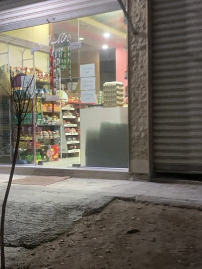 Looking through a store window, shelves are stocked with various products. There are signs displayed on the window and more merchandise is stacked on the shelves behind. Outside, the sidewalk is adjacent to a rough, textured wall. An awning partially covers the top of the window. A dark cable or wire extends into the foreground of the view.
