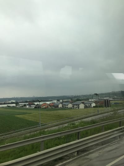 The image depicts an expansive, overcast landscape in Șcheia, Romania, observed through a reflective glass surface, indicated by indistinct reflections in the upper portion. The sky, occupying the upper two-thirds of the frame, is uniformly grey with heavy cloud cover. In the immediate foreground, a grey, multi-bar metal highway guardrail extends horizontally across the bottom quarter of the image, partially obscuring a segment of asphalt roadway with a white dividing line.

Beyond the guardrail, a strip of unkempt green vegetation separates the foreground from a multi-lane highway that curves subtly through the midground. Adjacent to the highway are distinct agricultural fields; a field with parallel rows of dark green crops is prominent on the left, while lighter green and less defined cultivated areas stretch towards the right. Scattered amidst and beyond these fields are various structures: a cluster of residential houses with multi-colored pitched roofs (orange, grey, brown) and light-toned facades, along with several larger, white, flat-roofed industrial or commercial buildings, predominantly on the left side. A utility pole featuring a solar panel array is centrally positioned within the midground.

In the distant background, a dense urban skyline, likely the city of Suceava, is silhouetted against the horizon, situated on an elevated landform. Architectural features, including a distinctive spire, are discernible within this urban profile. A body of water, possibly the Suceava River, is faintly visible on the far left horizon, reflecting the grey sky. No human figures are present in the scene.