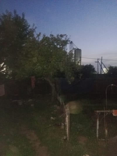 A grainy, dark image depicts a rural backyard scene at dusk. A large tree dominates the left side of the frame, its branches casting shadows. In the background, a tall concrete silo-like structure with a metal framework is visible, suggesting an agricultural or industrial setting. A power line extends across the sky. The ground is covered in grass and dirt, with some scattered debris. A metal table or stand is visible on the right side of the image, with an orange object on it. The sky is a deep blue, transitioning to a lighter shade towards the horizon, indicating the transition from day to night. The overall impression is one of a quiet, somewhat neglected outdoor space. The location is Atbasar, Kazakhstan.
