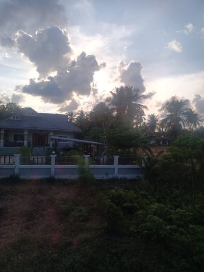 The image shows a residence in Hat Sai Ri, Thailand, observed from an elevated position. The house features a gray tiled roof and is situated behind a concrete fence with white pillars. Vegetation, including palm trees, surrounds the property. The foreground is dominated by thick foliage. The sky is partially overcast with sunlight breaking through the clouds.